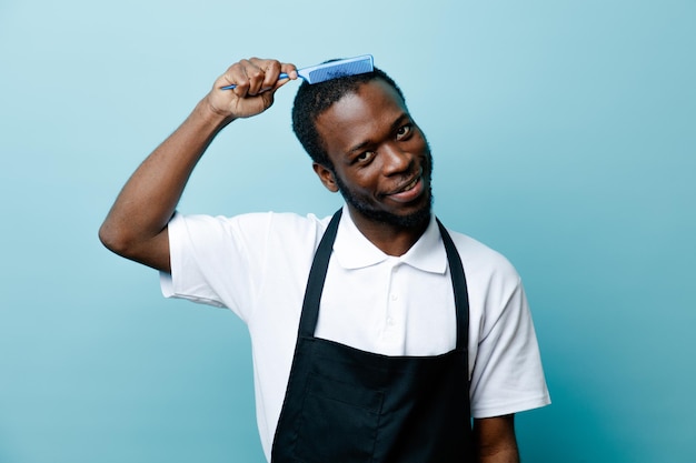 Blij haar kammen met kam jonge Afro-Amerikaanse kapper in uniform geïsoleerd op blauwe achtergrond