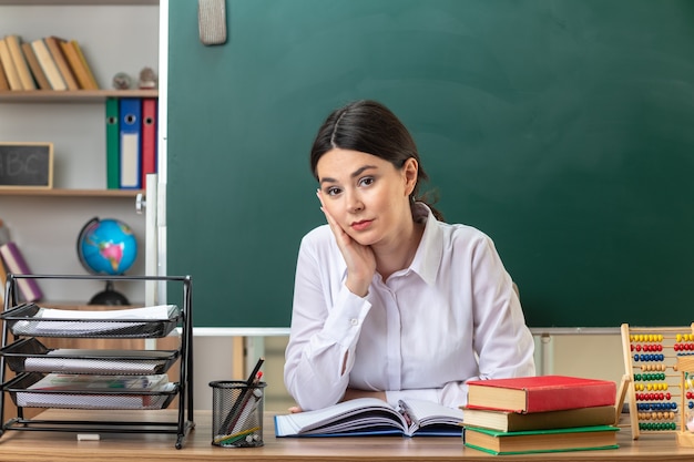 Blij hand op de wang te leggen jonge vrouwelijke leraar zittend aan tafel met schoolhulpmiddelen in de klas