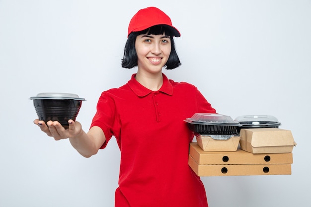 Blij jong kaukasisch bezorgmeisje met voedselcontainer en voedselverpakking op pizzadozen