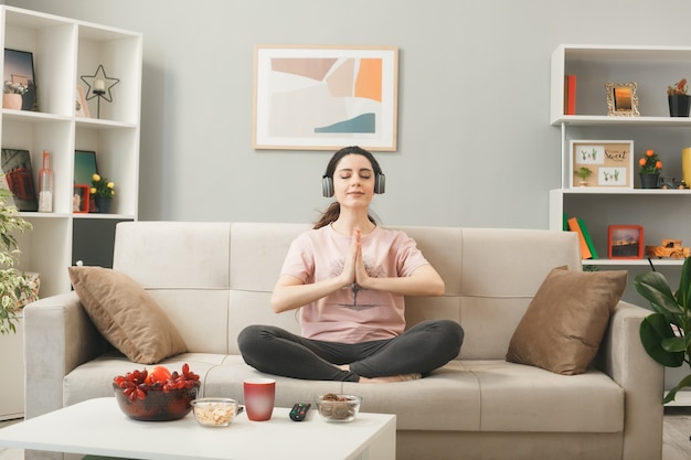 Blij jong meisje met een koptelefoon die yoga doet, zittend op de bank achter de salontafel in de woonkamer