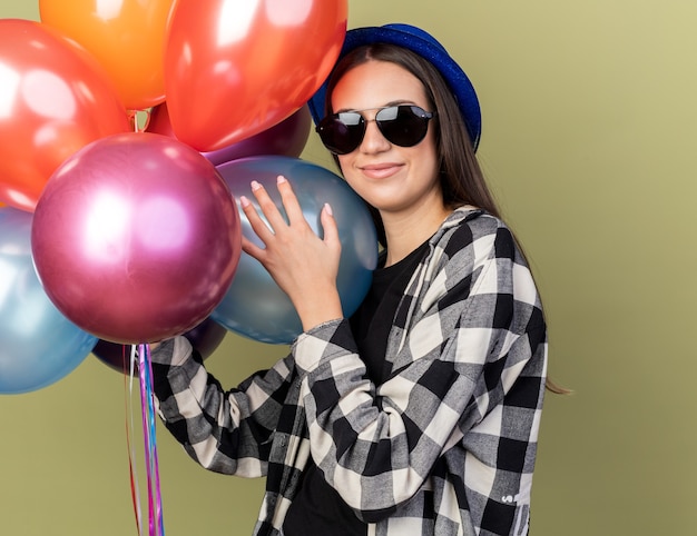 Blij jong mooi meisje met blauwe hoed met bril met ballonnen geïsoleerd op olijfgroene muur