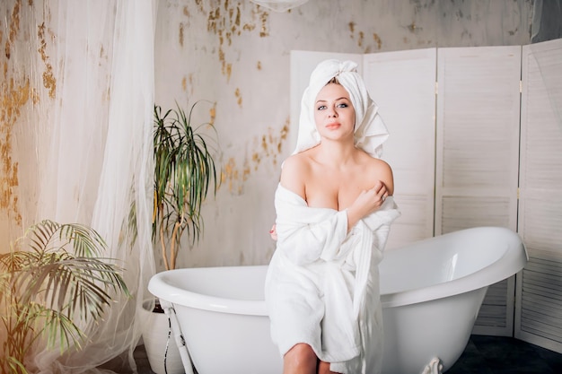 Blij jonge sexy vrouw in witte badjas met handdoek op hoofd kijken met plezier naar de camera, zittend aan de rand van de badkuip met blote schouders in luxe badkamer in de ochtend.