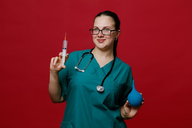 Blij jonge vrouwelijke arts dragen van een bril uniform en stethoscoop om haar nek kijken camera weergegeven: spuit en klysma geïsoleerd op rode achtergrond