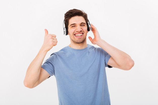 Blij man in casual t-shirt luisteren naar muziek via de koptelefoon en duim omhoog met een glimlach, geïsoleerd over witte muur