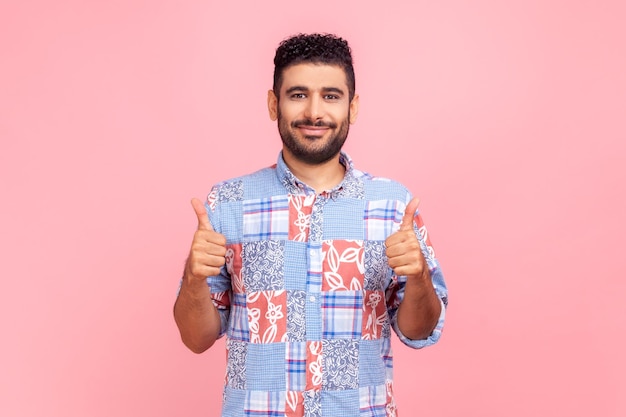 Blij man met baard in blauw shirt glimlachen naar de camera en gebaren duimen omhoog goedkeuring hand teken tevreden met resultaat Indoor studio shot geïsoleerd op roze achtergrond