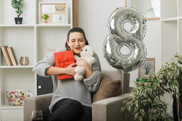 Blij met gesloten ogen mooi meisje op gelukkige vrouwendag met cadeau met teddybeer zittend op fauteuil in woonkamer