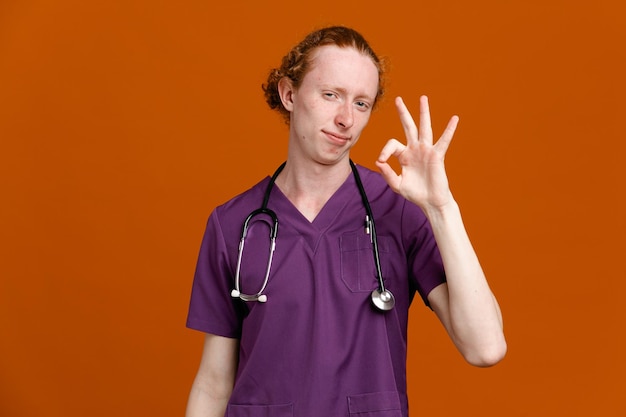Blij met goed gebaar jonge mannelijke arts in uniform met stethoscoop geïsoleerd op oranje achtergrond