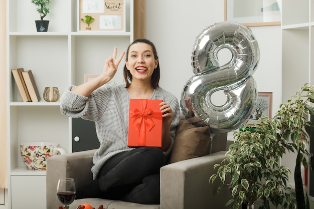 Blij met tong en vredesgebaar mooi meisje op gelukkige vrouwendag met cadeau zittend op fauteuil in woonkamer