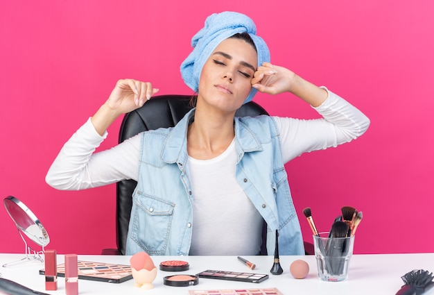 Blij mooie blanke vrouw met gewikkeld haar in een handdoek zittend aan tafel met make-up tools die handen opsteken geïsoleerd op roze muur met kopieerruimte