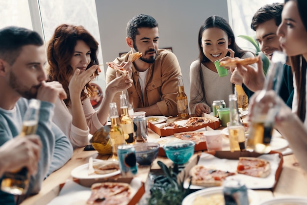 Blij om in de buurt te zijn. Groep jongeren in vrijetijdskleding die pizza eet en glimlacht terwijl ze binnen een etentje hebben