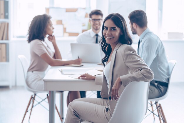 Blij om in team te zijn. Mooie vrolijke vrouw die met een glimlach naar de camera kijkt terwijl ze met haar collega's aan de kantoortafel zit