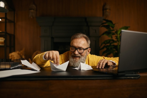 Blij opgewonden volwassen grijsharige man die met een papieren bootje speelt terwijl hij op de werkplek in het thuiskantoor zit. Dromen over toekomstige reizen of vakantie op pensioenconcept