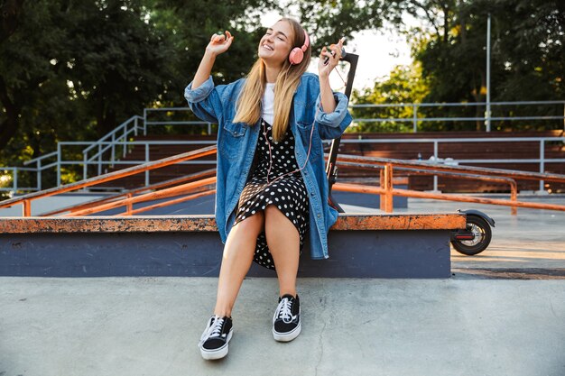 blij optimistisch jong tienermeisje buiten in park luisteren muziek met koptelefoon met mobiele telefoon dansen.