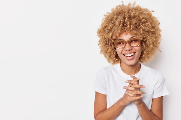 Blij positieve vrouw houdt handen bij elkaar glimlacht in grote lijnen voelt zich tevreden herinnert zich grappig verhaal gebeurde met haar gekleed in t-shirt geïsoleerd op witte achtergrond met kopie ruimte voor uw promotie