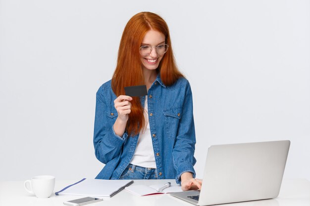 Blij, tevreden ontspannen schattige roodharige vrouw die een bestelling plaatst op haar werk, online betaalt, creditcardgegevens invoert, factuurgegevens invoert, laptop gebruikt om te kopen, staand wit