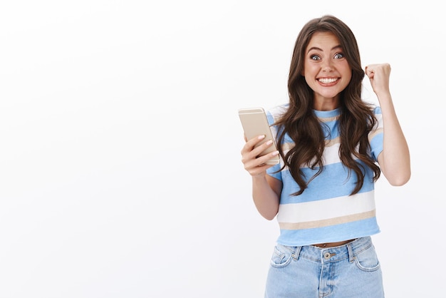 Blij vrolijk in t-shirt en spijkerbroek glimlachend triomfantelijk, ontvang fantastisch goed nieuws, houd smartphone vast, vuist pomp vreugdevol