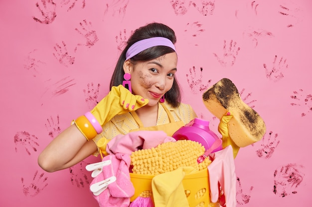 Blije brunette Aziatische huisvrouw beschermt huis tegen vuil toont vuile spons na het afvegen van meubels draagt hoofdband latex handschoenen poseert in de buurt van wasmand gebruikt schoonmaakspullen heeft gezicht gevlekt