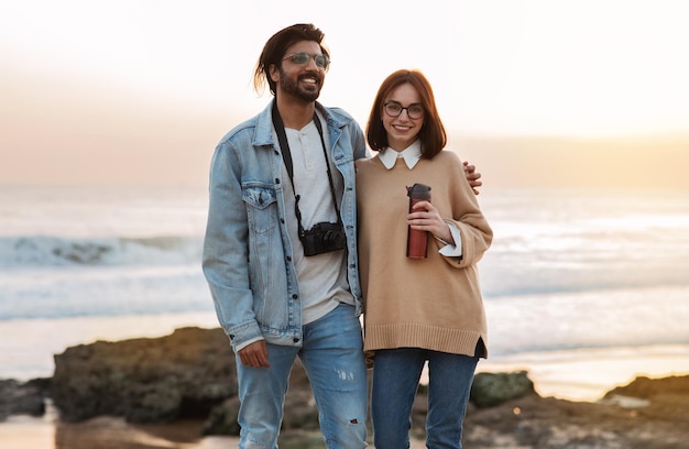 Blije duizendjarige indiase man die een blanke dame met een thermosfles knuffelt, geniet van oceaanwandelingen op het strand buiten