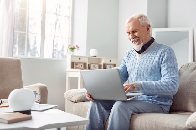 Blije gebruiker. Vrolijke oudere man zittend op de bank in de woonkamer en met behulp van zijn laptop, een bericht typen aan zijn vrienden