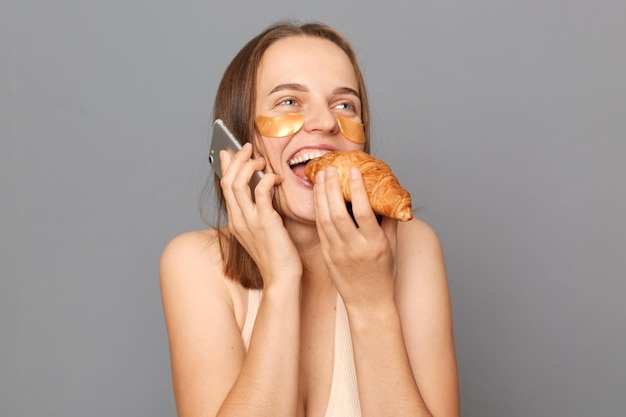 Blije hongerige jonge vrouw met bruin haar poseren geïsoleerd over grijze achtergrond staande met gepatchte onder ogen croissant etend pratend op mobiele telefoon