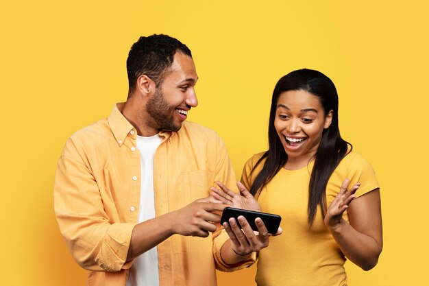 Blije jonge afro-amerikaanse man die telefoon laat zien aan geschokte vrouw geïsoleerd op gele achtergrondstudio