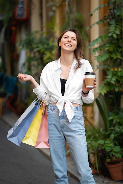 Blije jonge blanke dame met veel tassen met aankopen geniet van een winkeltour in een nieuwe stad met een kopje koffie