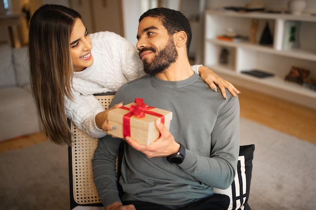 Blije jonge blanke vrouw feliciteert man uit het Midden-Oosten geeft doos met cadeau in kamerinterieur