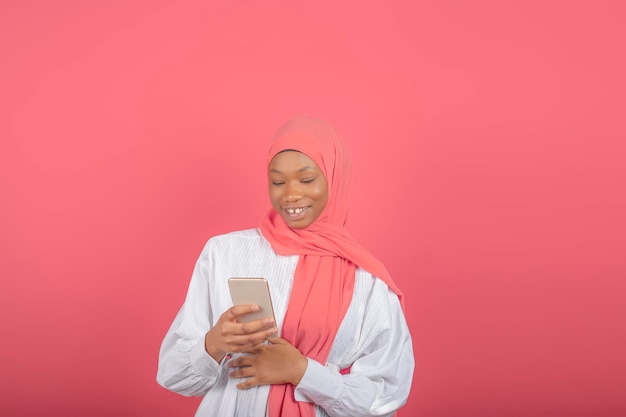 Blije jonge moslimvrouw gebruikt smartphone om te socializen, reageert in online chat, plaatst iets op sociale media, draagt roze sluier en wit overhemd, geïsoleerd op roze achtergrond.