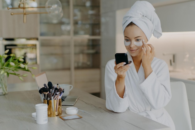 Blije jonge vrouw draagt badjas en handdoek gewikkeld op het hoofd zorgt voor de huid onder de ogen brengt schoonheidspleisters aan kijkt naar zichzelf in de spiegel zit aan tafel met laptop en cosmetische hulpmiddelen poseert thuis