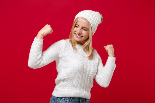 Foto blije jonge vrouw in witte sweaterhoed die op rood muur achtergrondstudioportret wordt geïsoleerd. gezonde mode lifestyle mensen emoties koude seizoen concept. mock-up kopie ruimte. gebalde vuisten als winnaar.