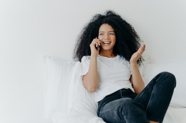 Blije, krullende, etnische dame heeft telefoongesprekken, gebaren en lacht van vreugde, kijkt ergens opzij