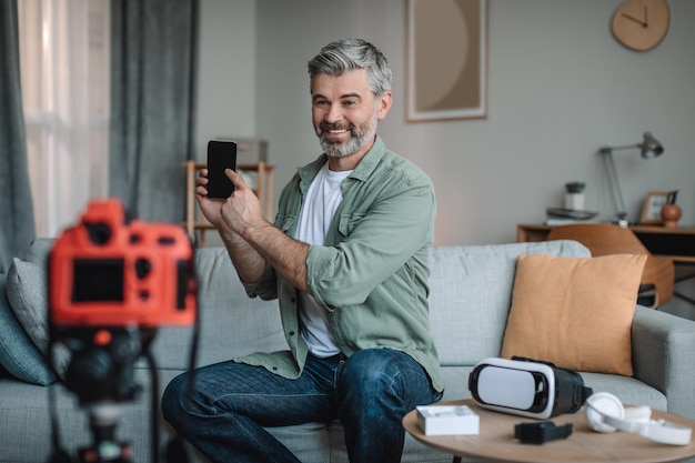 Foto blije oude europese man neemt videobeoordeling op apparaat op en toont smartphone met leeg scherm in de kamer