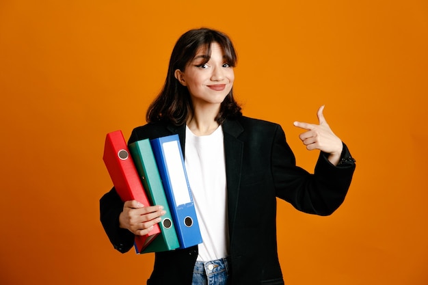 Blije punten aan de zijkant met mappen jonge mooie vrouw met zwarte jas geïsoleerd op een oranje achtergrond