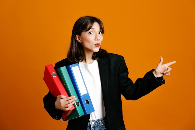 Blije punten aan de zijkant met mappen jonge mooie vrouw met zwarte jas geïsoleerd op een oranje achtergrond