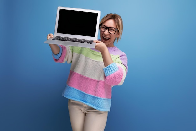 Blije succesvolle blonde studente in een gestreepte trui demonstreert een model van een laptop op een