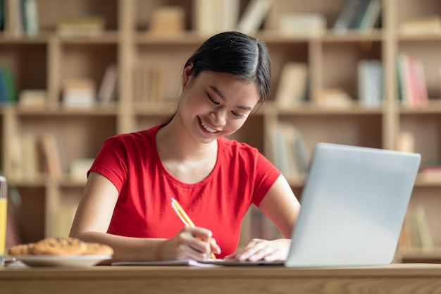 Blije tiener Aziatische vrouw maakt aantekeningen aan tafel met laptopstudie in het interieur van de woonkamer