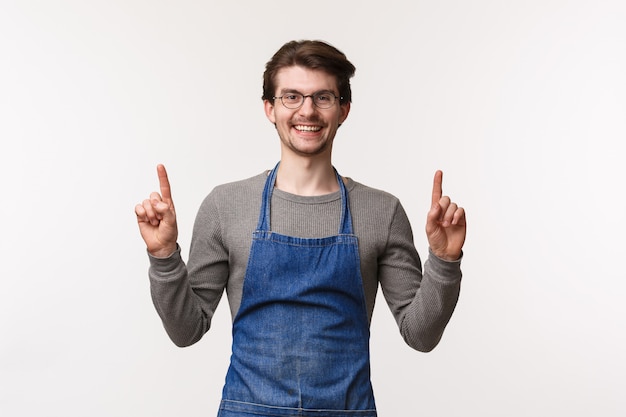 Blije, vriendelijk ogende barman nodigt zijn bezoek uit, wijst met zijn vingers omhoog en lacht vrolijk naar de camera terwijl hij een schort draagt op het werk, koffie zet voor de klant, stoelen boven toont,