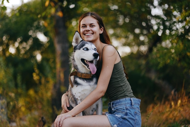 Blije vrouw met een husky-rashond die lacht terwijl ze in de natuur zit tijdens een wandeling met een hond aan de lijn herfstlandschap op de achtergrond