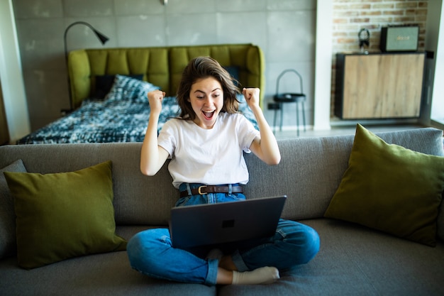 Blije vrouw met winstgebaar die laptop op de bank met behulp van