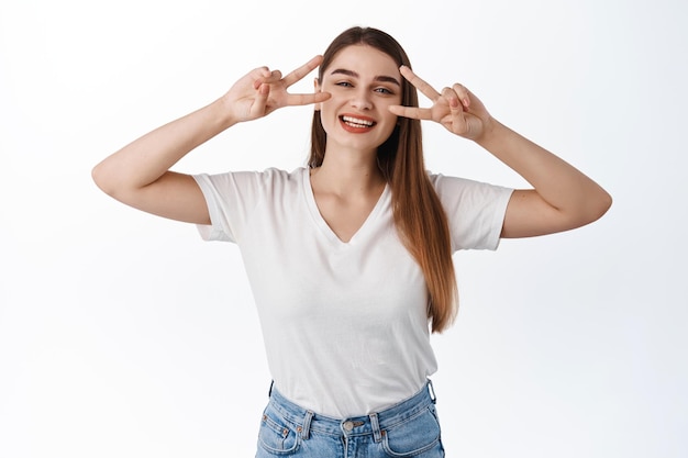 Blijf positief. glimlachend mooi meisje toont vrede v-teken en kijkt gelukkig naar de camera, drukt heldere vreugdevolle emoties uit, geniet van de lente, staande in t-shirt op witte achtergrond