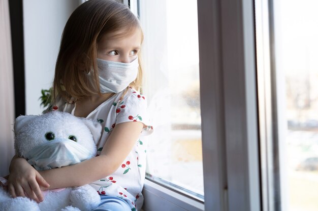Blijf thuis quarantaine coronavirus pandemie preventie. verdrietig kind en zijn teddybeer beide in beschermende medische maskers zit op de vensterbank en kijkt uit het raam