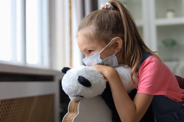 Blijf thuis quarantaine coronavirus pandemie preventie. Verdrietig kind met beschermend medisch masker en haar pandabeer zitten op een hobbelpaard en kijken uit het raam.