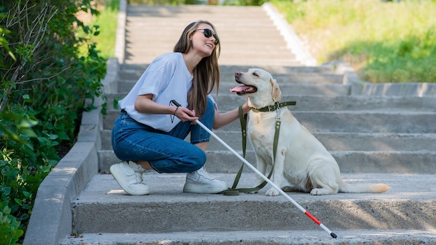 Blinde jonge blonde die een geleidehond aait tijdens een wandeling in het park vrouw met taststok bij de trap