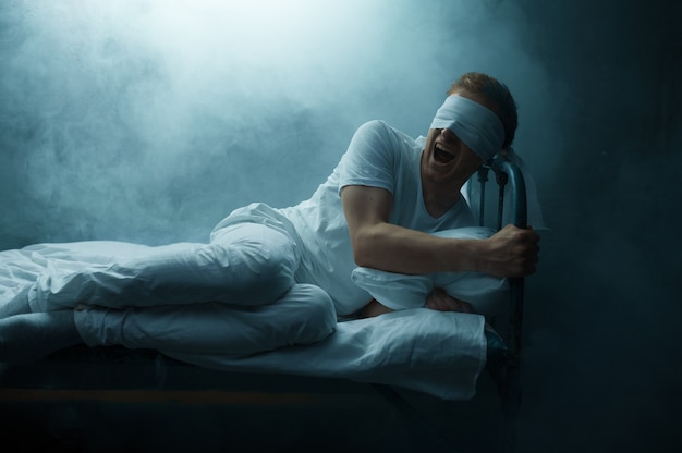 Blindfolded crazy man sitting in bed, dark room.. psychedelic male person having problems every night, depression and stress, sadness, psychiatry hospital