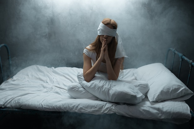 Donna pazza bendata seduta a letto, camera oscura .. persona di sesso femminile psichedelica che ha problemi ogni notte, depressione e stress, tristezza, ospedale psichiatrico