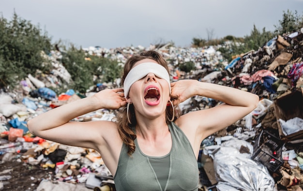 Женщина-волонтер с завязанными глазами кричит от бессилия на свалке пластикового мусора. День Земли и экология.