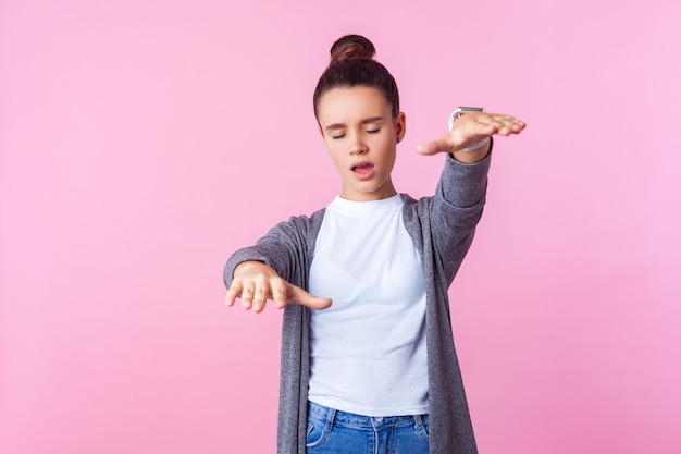 Blindness. Portrait of brunette teenage girl with bun hairstyle in casual clothes standing with closed eyes, disoriented reaching out hands to find way. indoor studio shot isolated on pink background