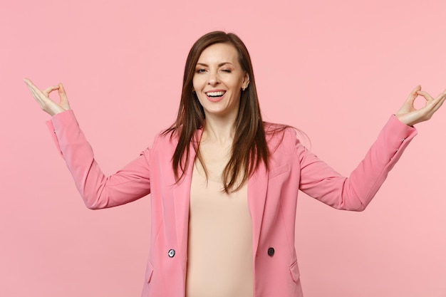 Blinking cheerful young woman in jacket hold hands in yoga gesture relaxing meditating isolated on pastel pink wall background in studio. People sincere emotions lifestyle concept. Mock up copy space.