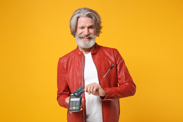 Blinking elderly gray-haired mustache bearded man isolated on yellow background. People lifestyle concept. Mock up copy space. Hold modern bank payment terminal process acquire credit card payments.