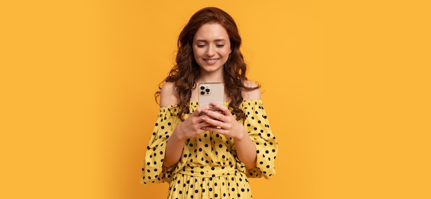 Blissful smiling red head woman holding mobile phone in yellow summer dress posing on yellow